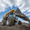 Yellow skid steer loader dumping dirt on rocky terrain with cloudy sky