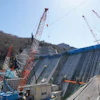 Large concrete dam under construction with red and white cranes on mountainside