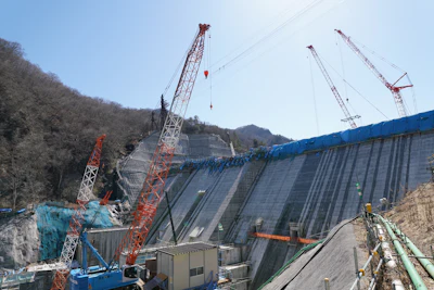 Large concrete dam under construction with red and white cranes on mountainside