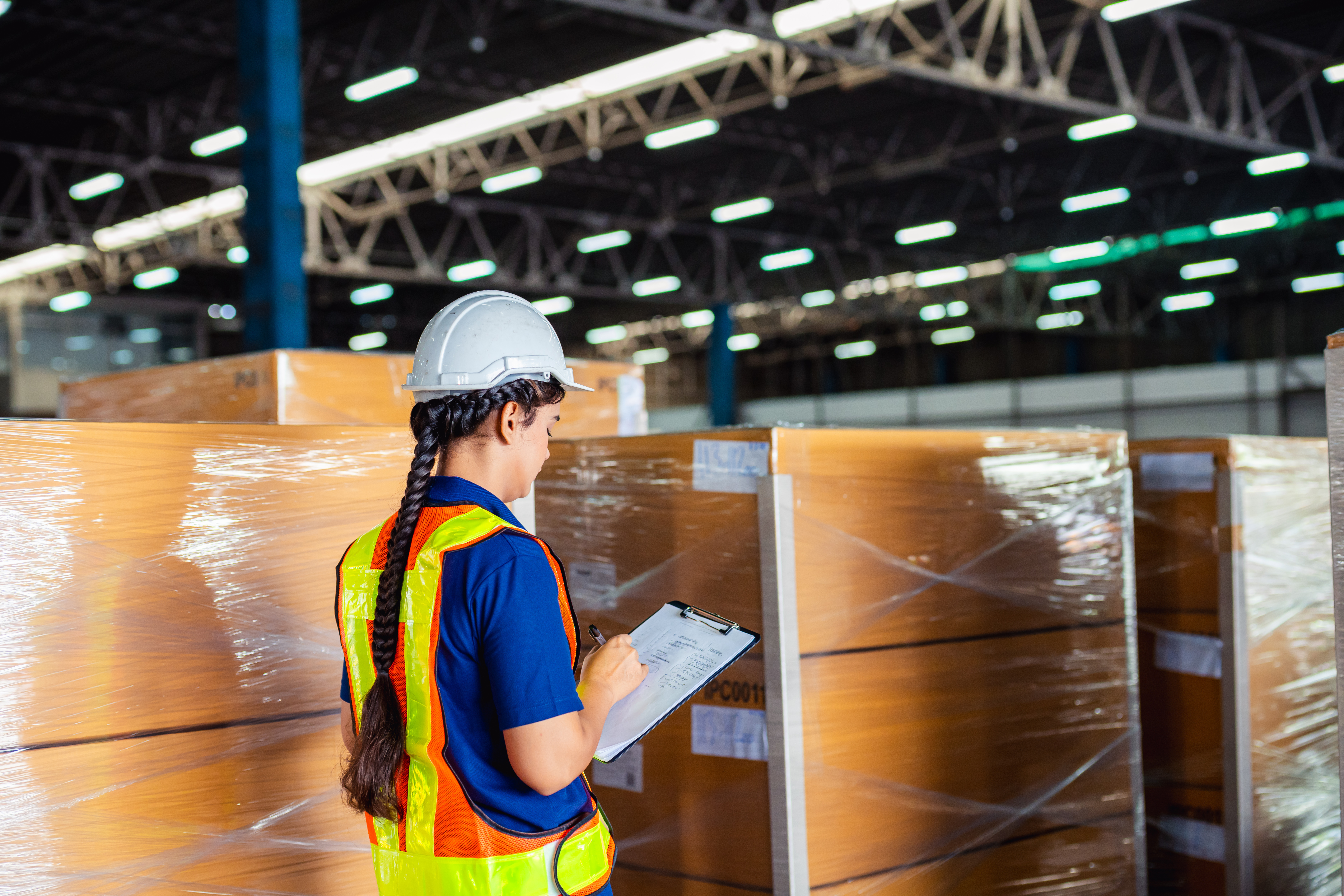 Warehouse worker in safety vest and hard hat reviewing clipboard near stacked wrapped pallets