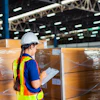 Warehouse worker in safety vest and hard hat reviewing clipboard near stacked wrapped pallets