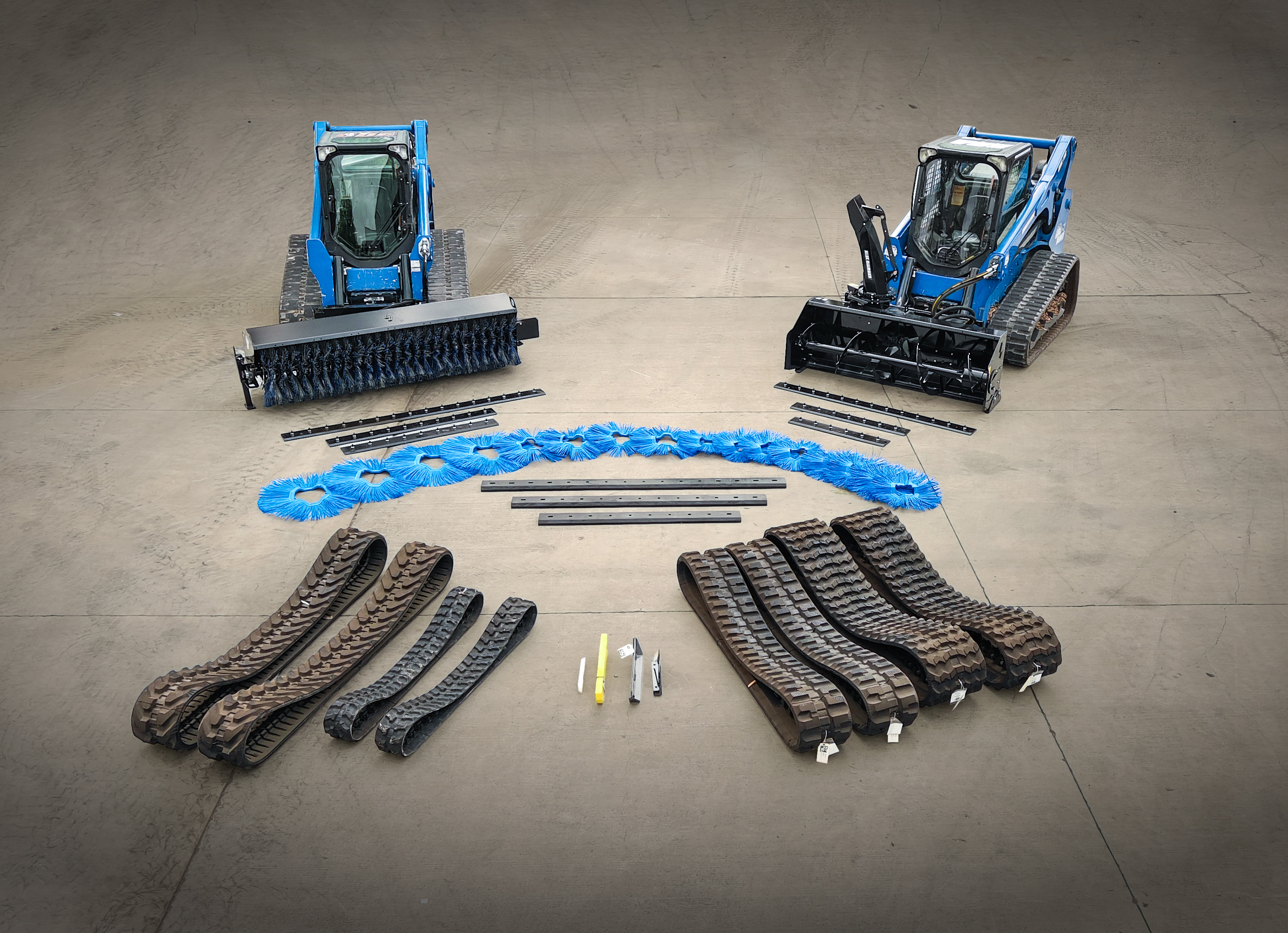 Two blue tracked machines with rubber tracks and attachments on concrete surface