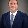 Professional businessman in navy suit and blue tie, smiling against gray background