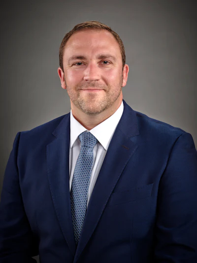 Professional businessman in navy suit and blue tie, smiling against gray background