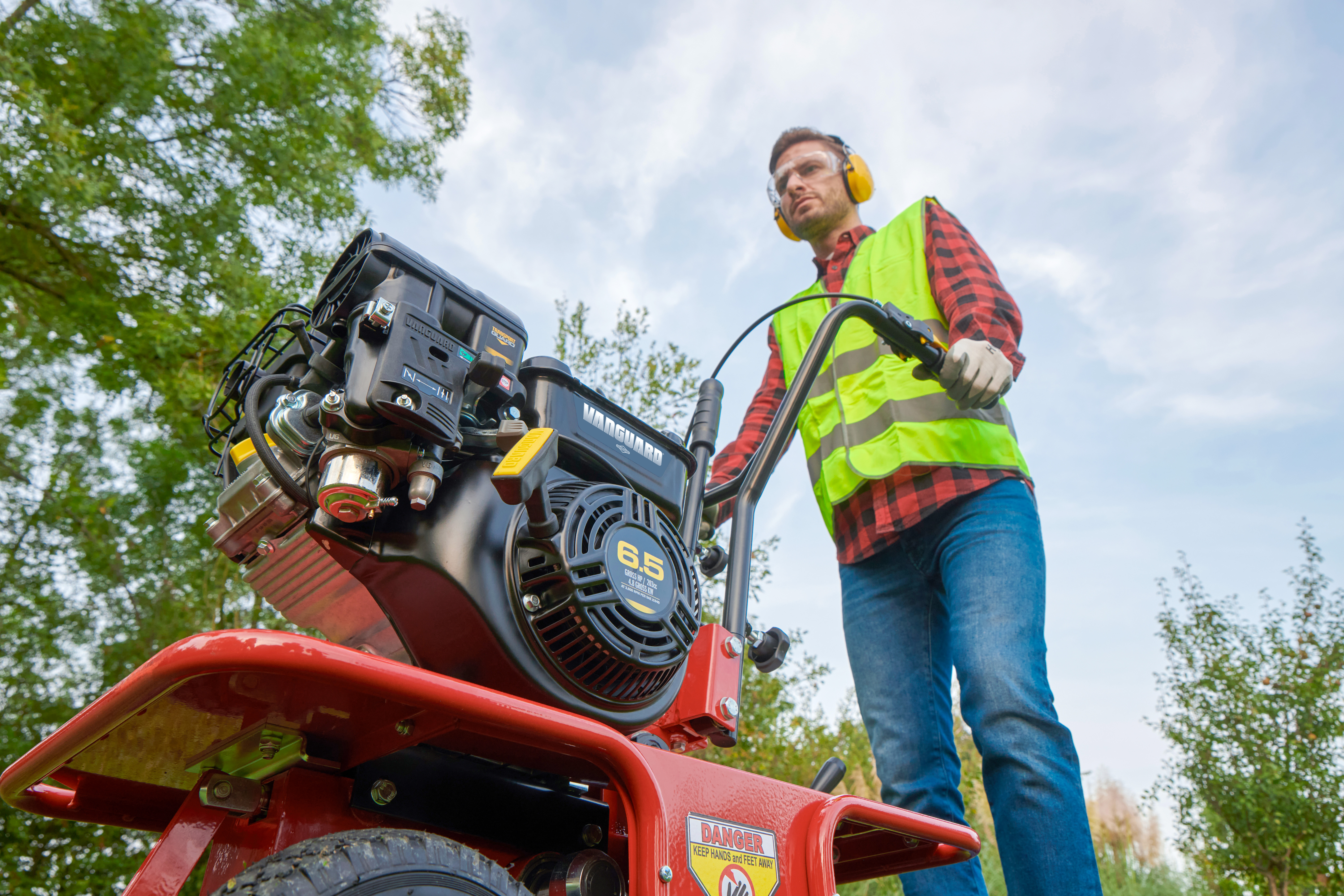 Worker in safety vest operating a red power tiller outdoors with trees in background