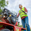 Worker in safety vest operating a red power tiller outdoors with trees in background