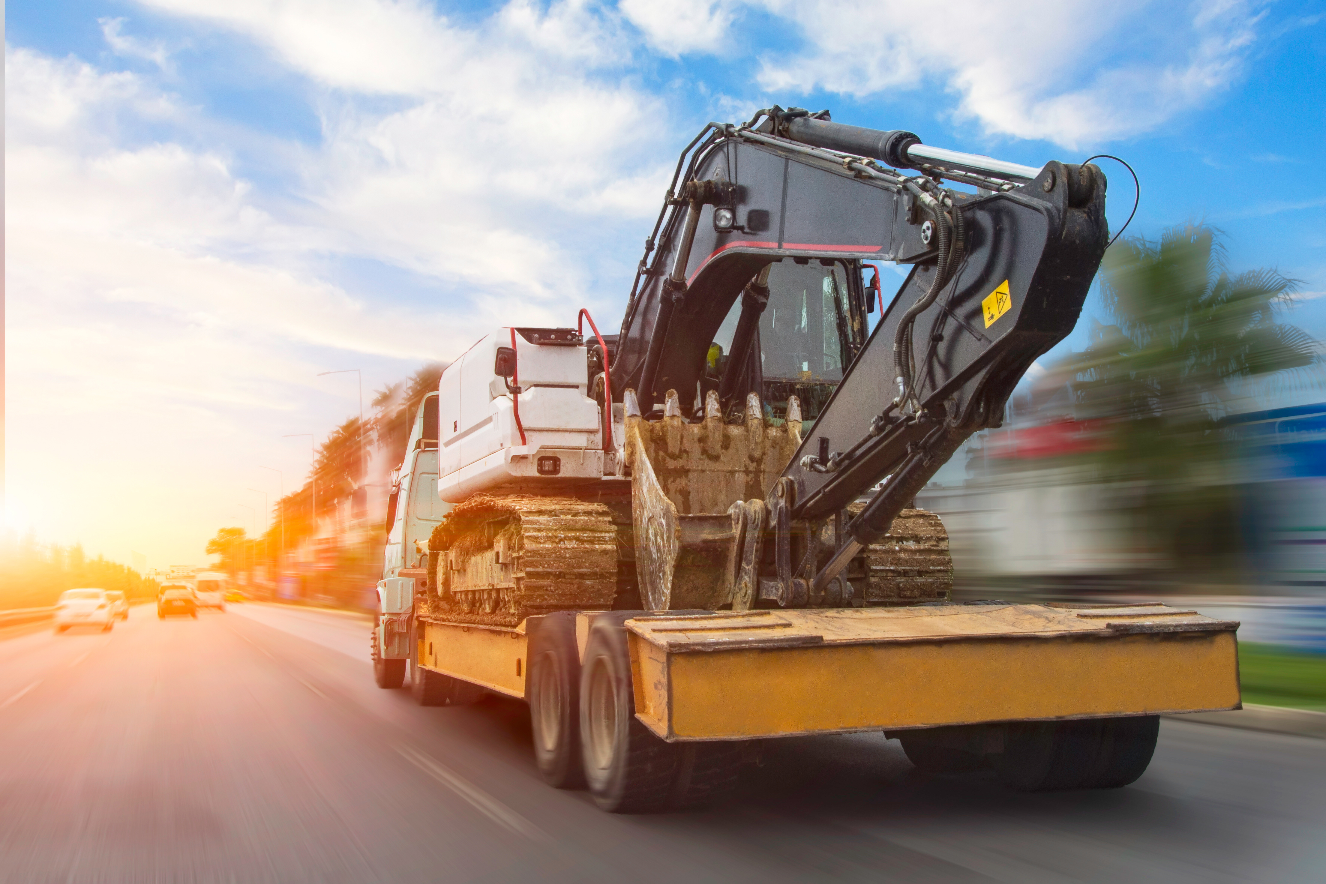 Yellow flatbed truck transporting gray excavator on highway at sunset