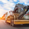 Yellow flatbed truck transporting gray excavator on highway at sunset