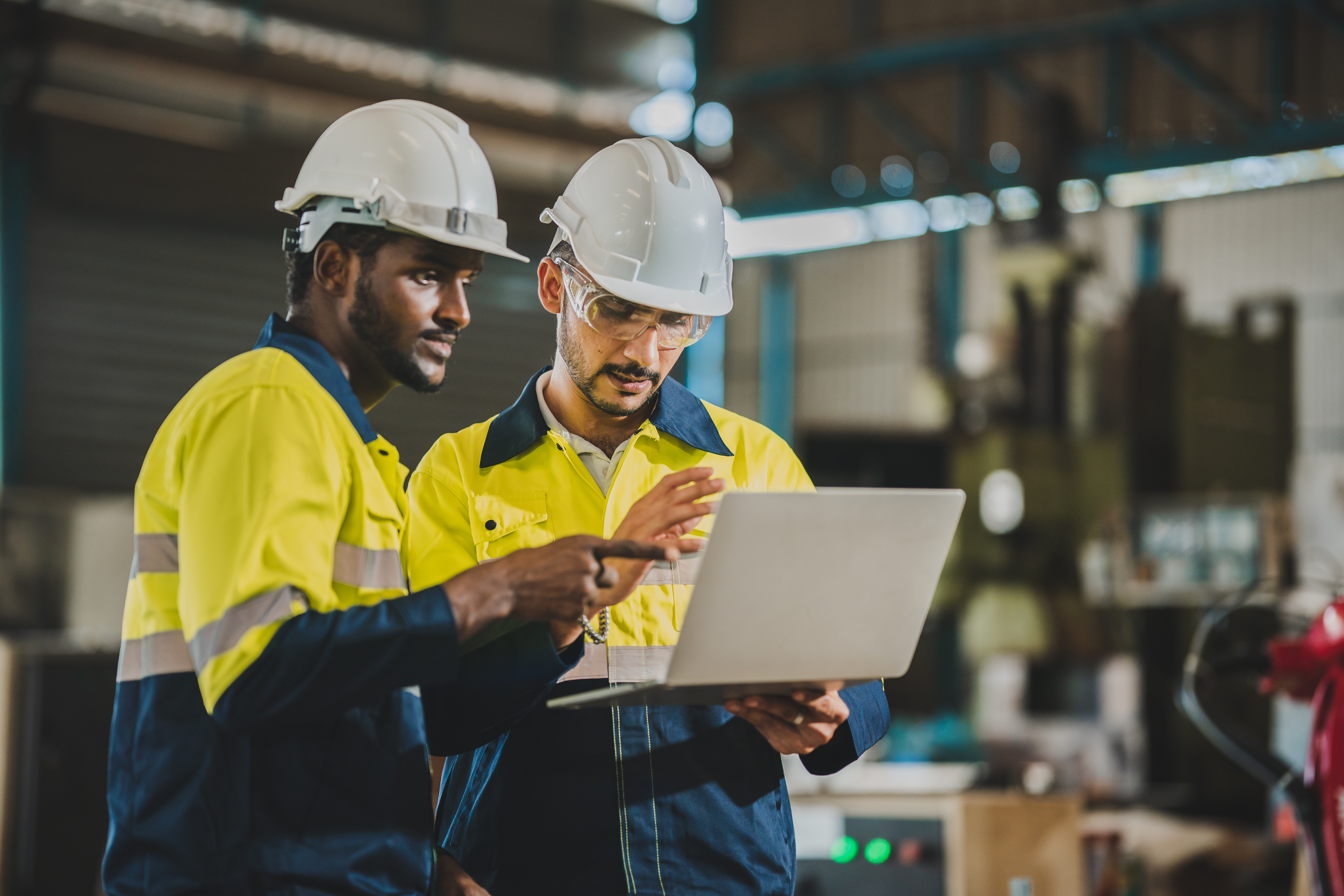 Two construction workers in safety gear reviewing laptop in industrial facility
