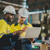 Two construction workers in safety gear reviewing laptop in industrial facility