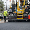 Road paving machine laying fresh asphalt on urban street with worker in safety vest