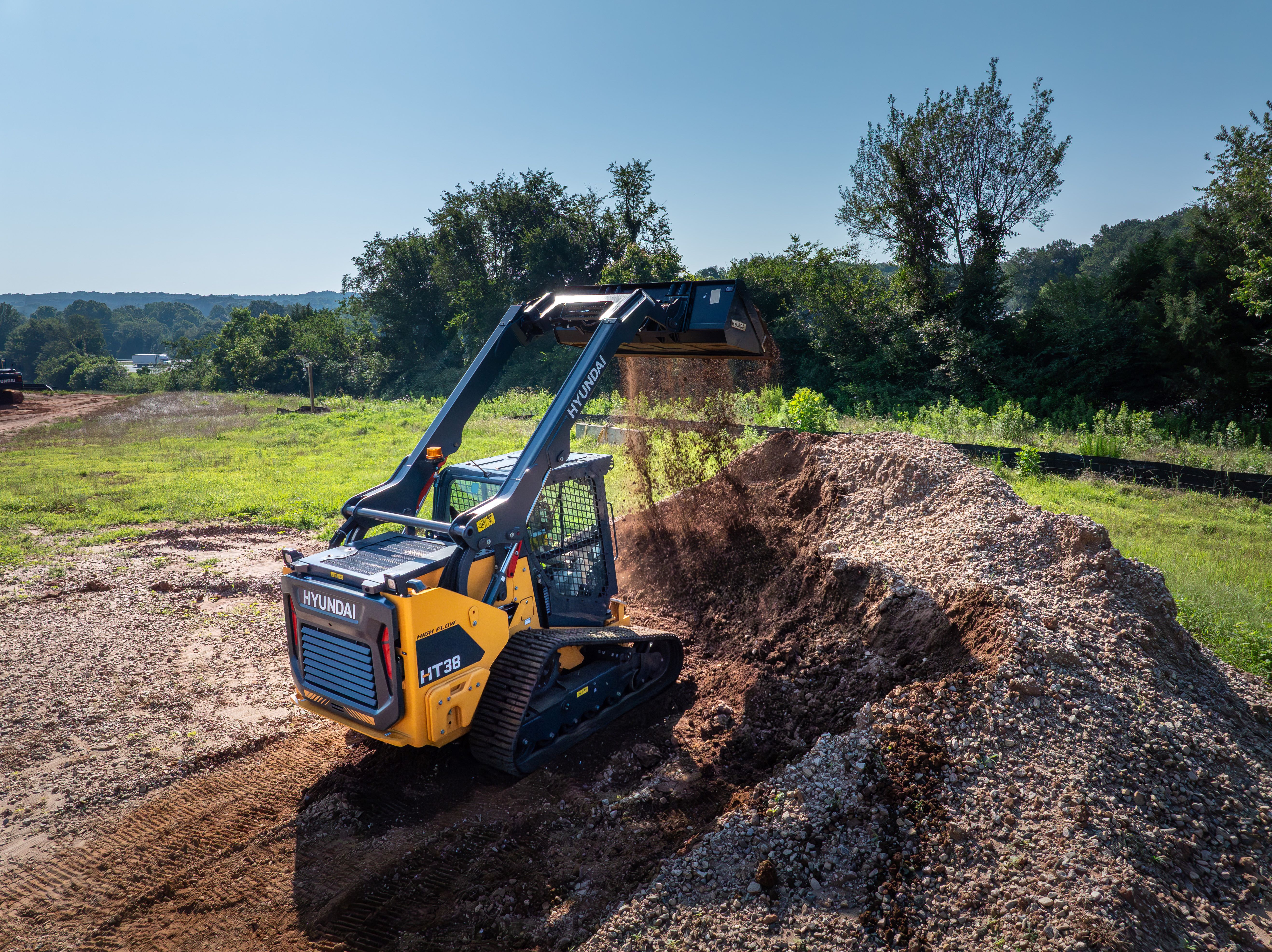 Yellow Hyundai tracked skid steer loader moving dirt on a construction site with green field background