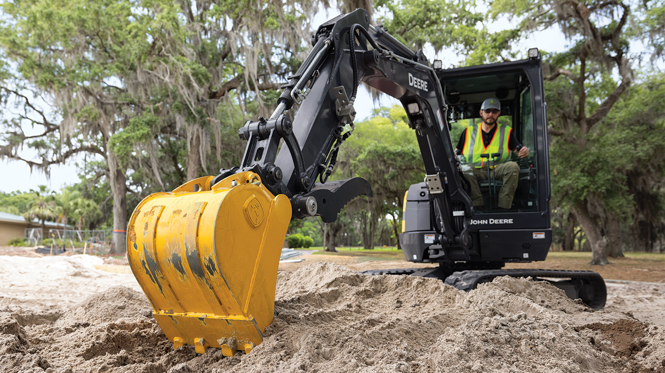 John Deere excavator with yellow bucket digging in sandy terrain near trees