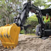 John Deere excavator with yellow bucket digging in sandy terrain near trees