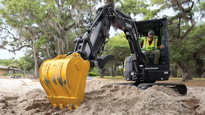 John Deere excavator with yellow bucket digging in sandy terrain near trees