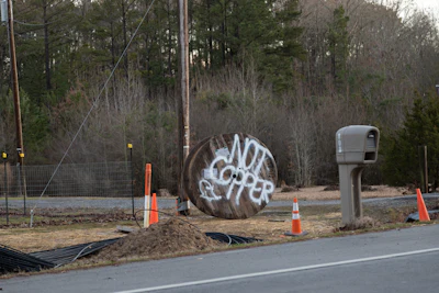 Construction site with traffic cones, utility pole, and graffitied metal cylinder near wooded area