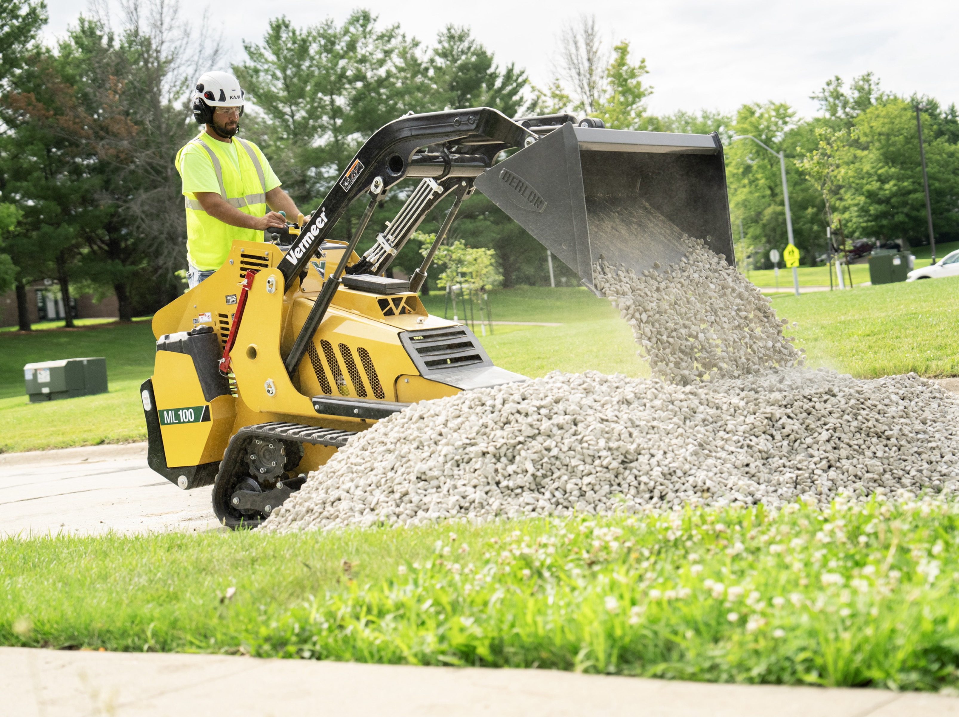 Yellow mini skid steer loader dumping gravel on grass near trees