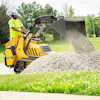Yellow mini skid steer loader dumping gravel on grass near trees