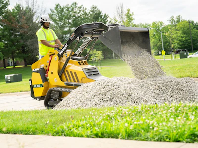 Yellow mini skid steer loader dumping gravel on grass near trees