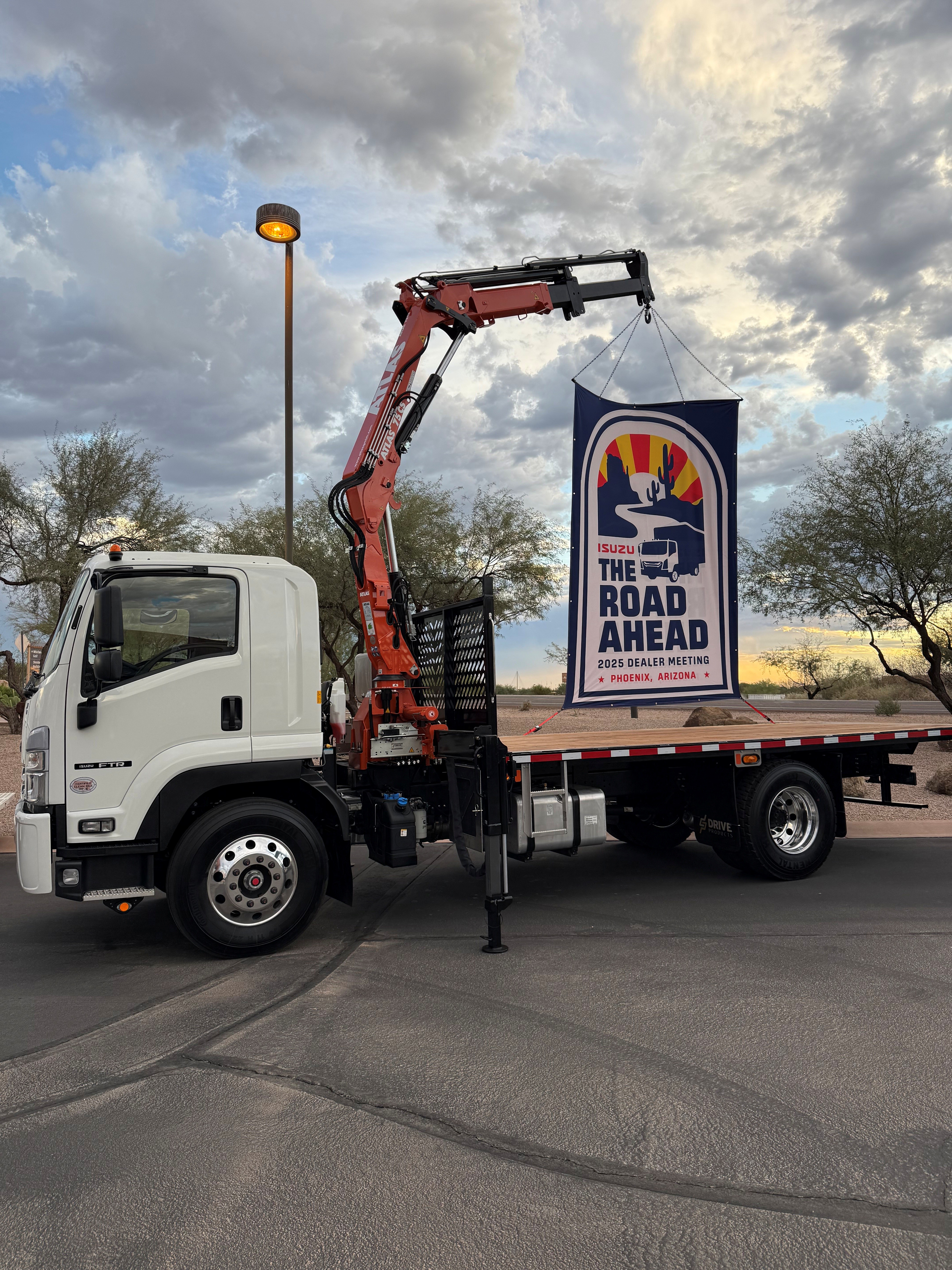 White Isuzu truck with crane displaying Road Ahead Dealer Meeting banner in parking lot