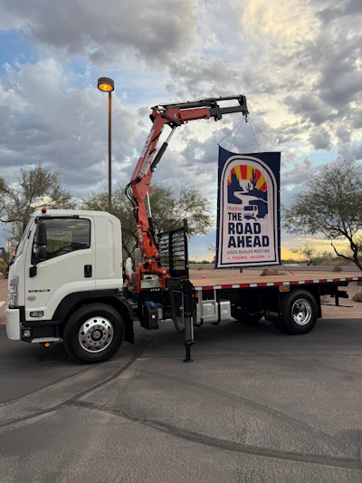 White Isuzu truck with crane displaying Road Ahead Dealer Meeting banner in parking lot