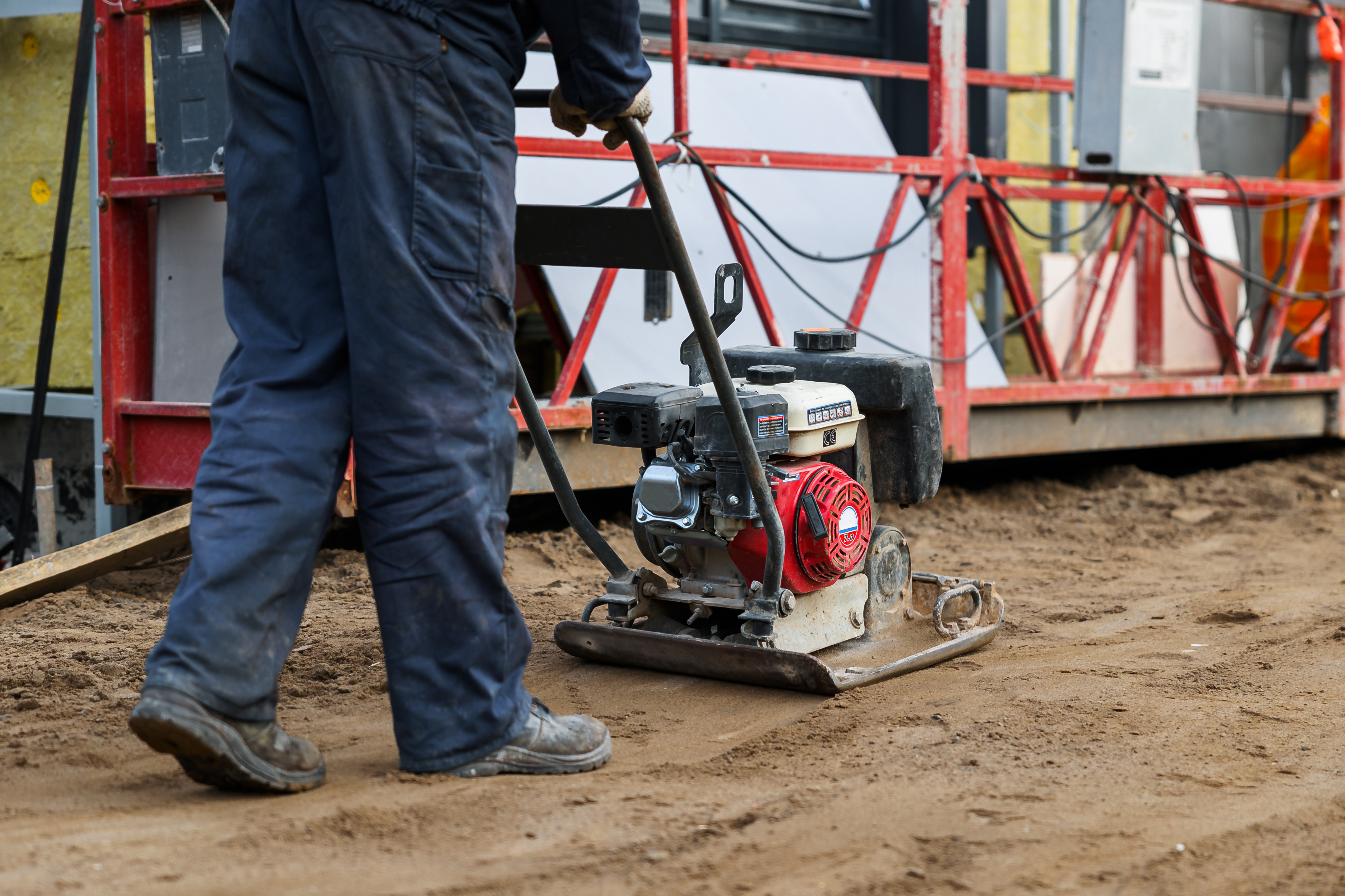 Construction worker operating plate compactor on dirt site with scaffolding