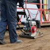 Construction worker operating plate compactor on dirt site with scaffolding