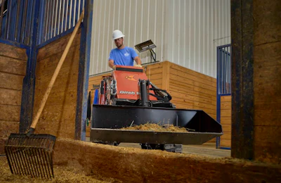 Worker operating red Ditch Witch machine in wooden barn stall with straw bedding