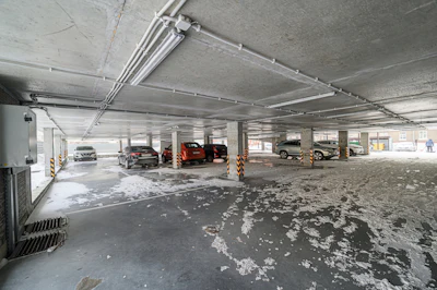 Underground parking garage with several vehicles parked near concrete pillars and exposed ceiling pipes