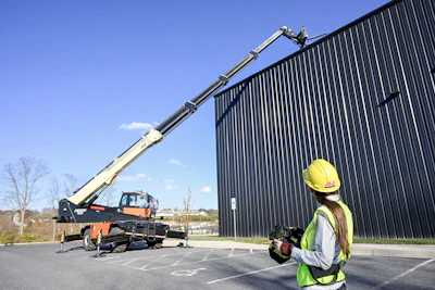 Construction worker operating mobile crane near metal warehouse building