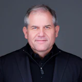 Professional headshot of a mature businessman in black attire against gray background