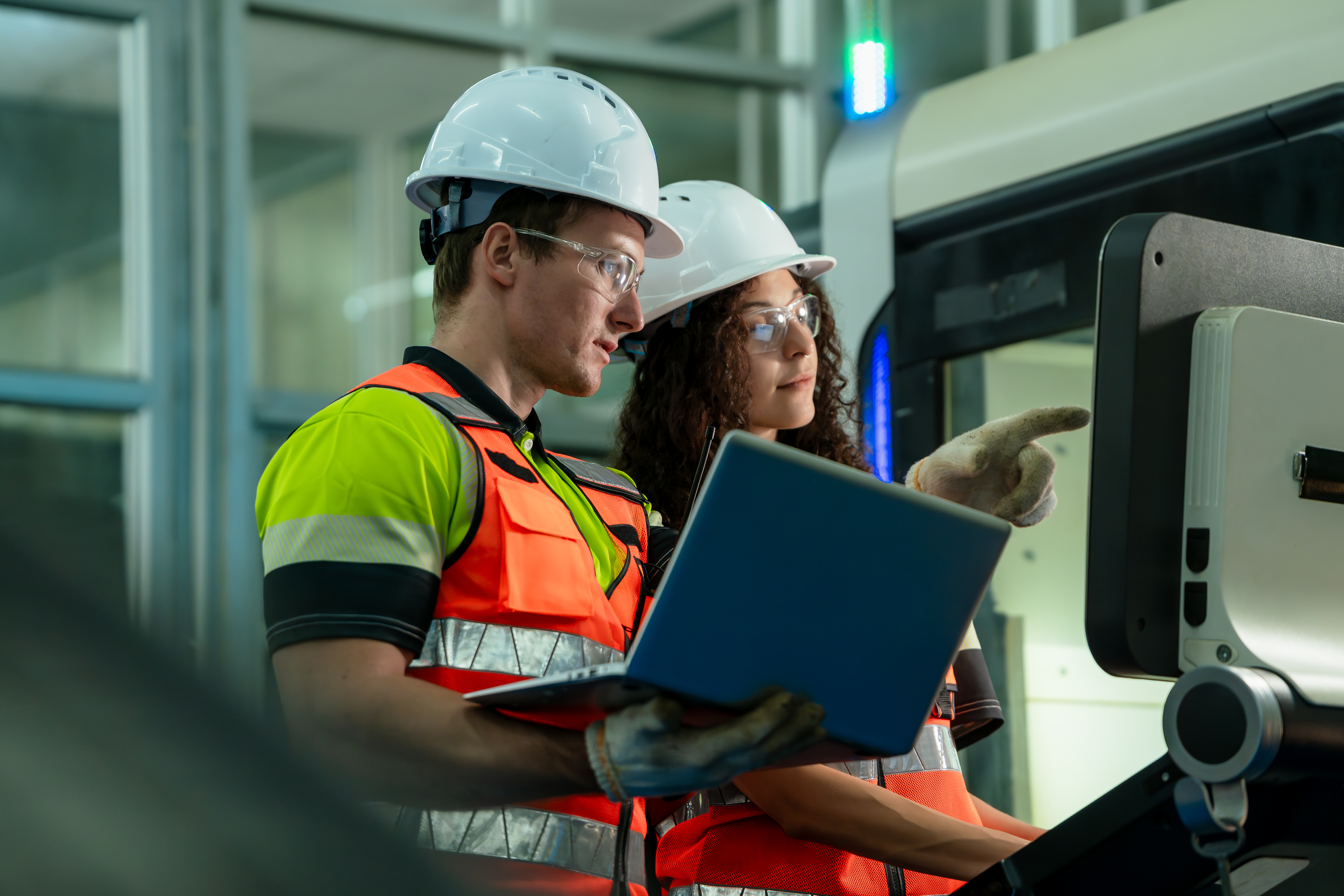 Two industrial workers in safety gear reviewing data on laptop near machinery