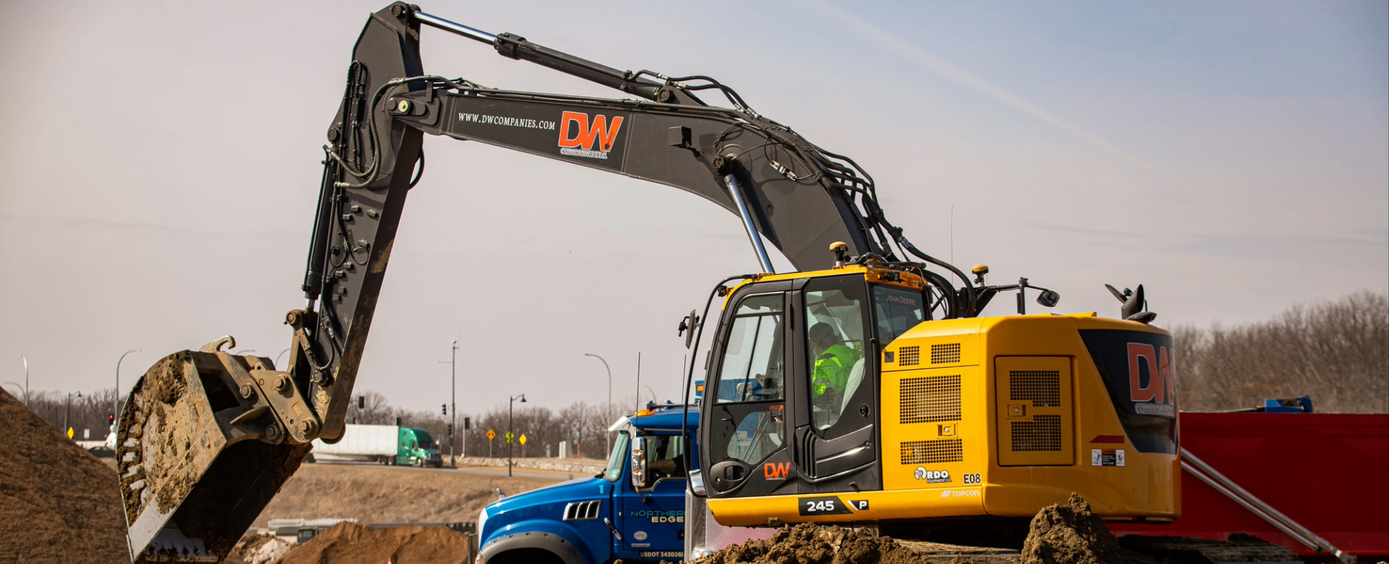 Yellow excavator and blue truck working on construction site with dirt mounds