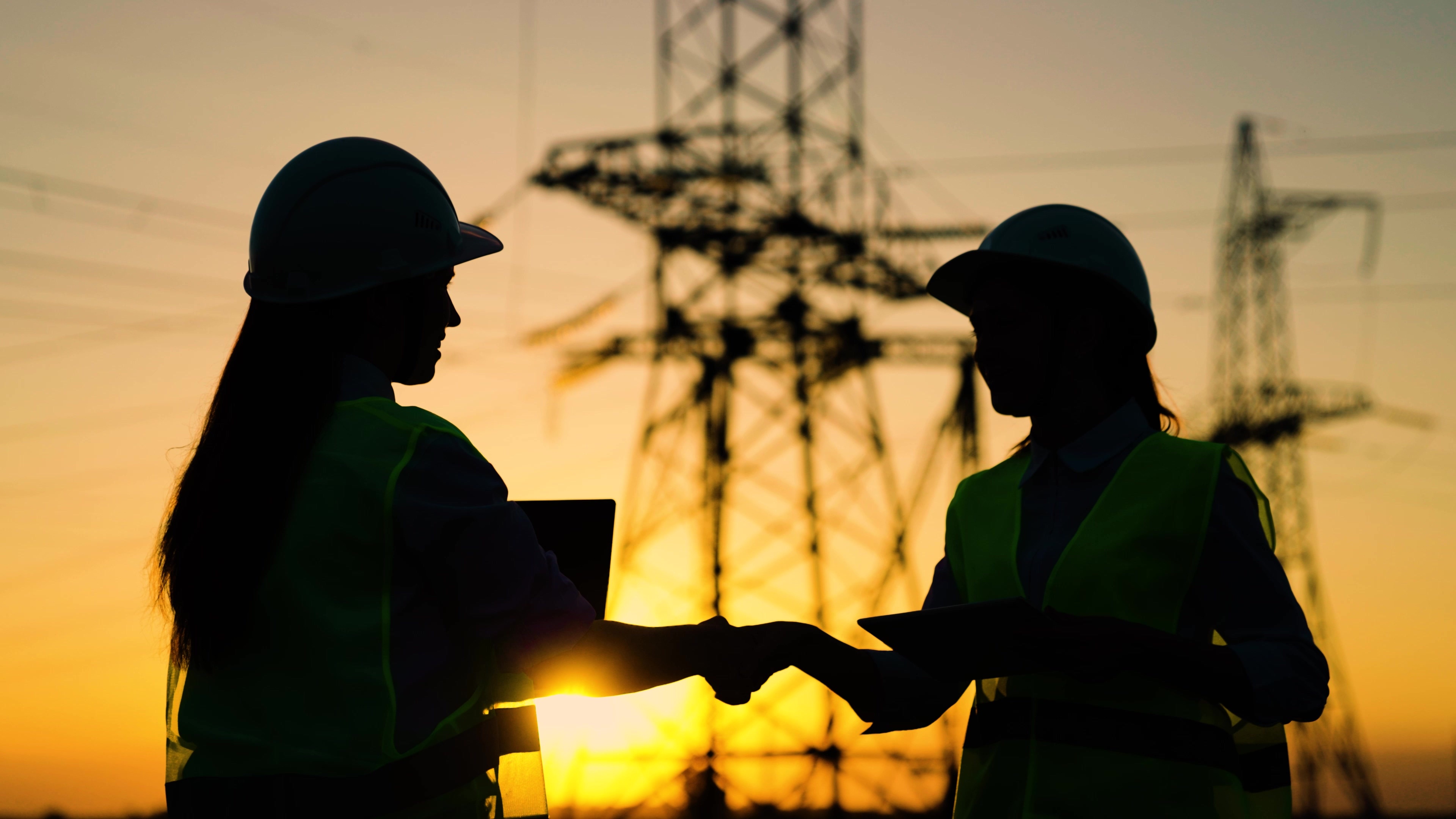 Two construction workers shaking hands at sunset near electrical towers