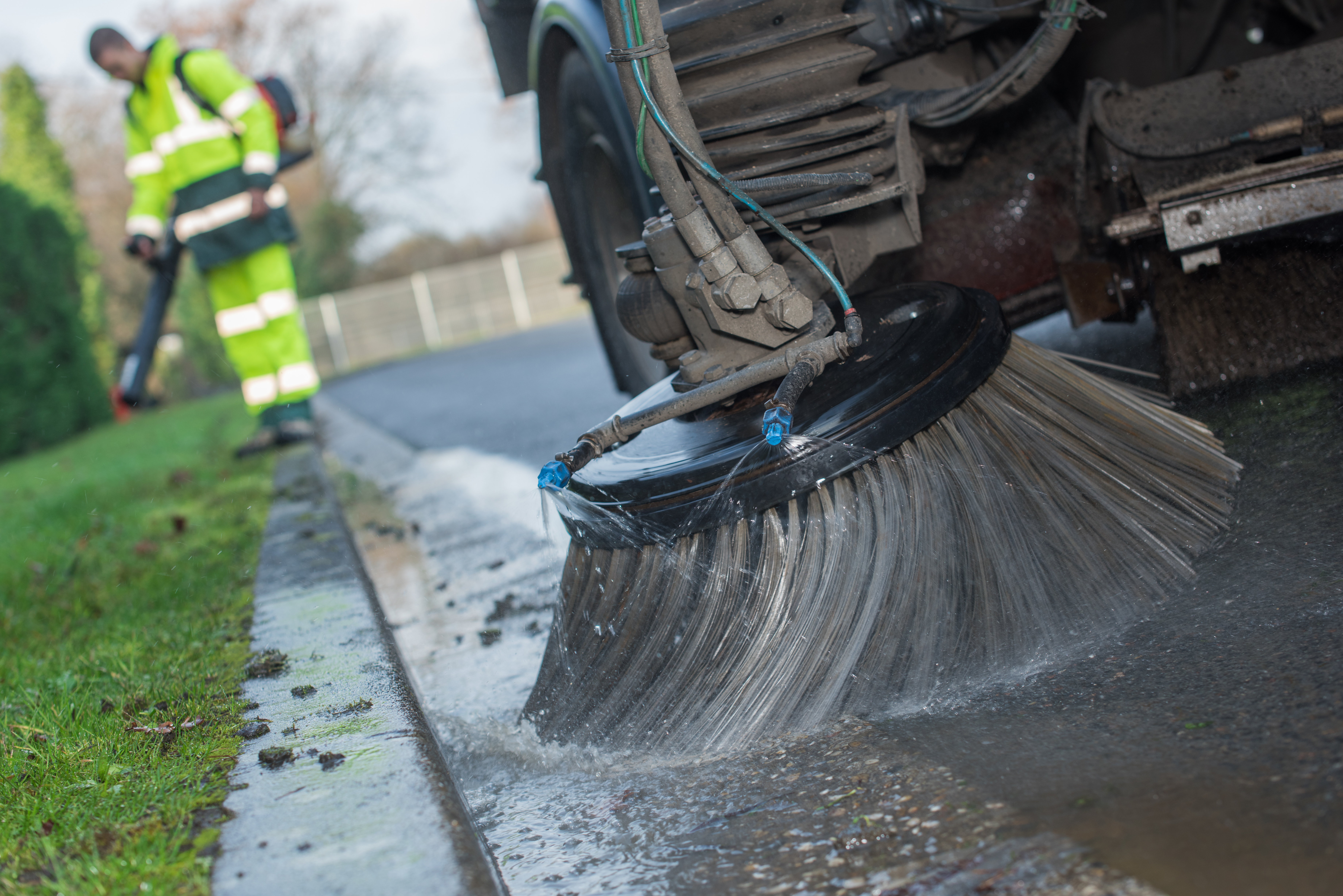 Street sweeper truck cleaning curb with large rotating brush, worker in high-visibility vest nearby