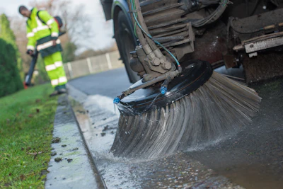 Street sweeper truck cleaning curb with large rotating brush, worker in high-visibility vest nearby