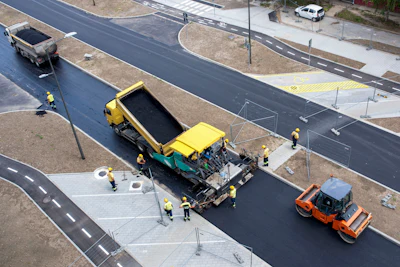 Road construction site with yellow paver, orange roller, and workers laying fresh asphalt