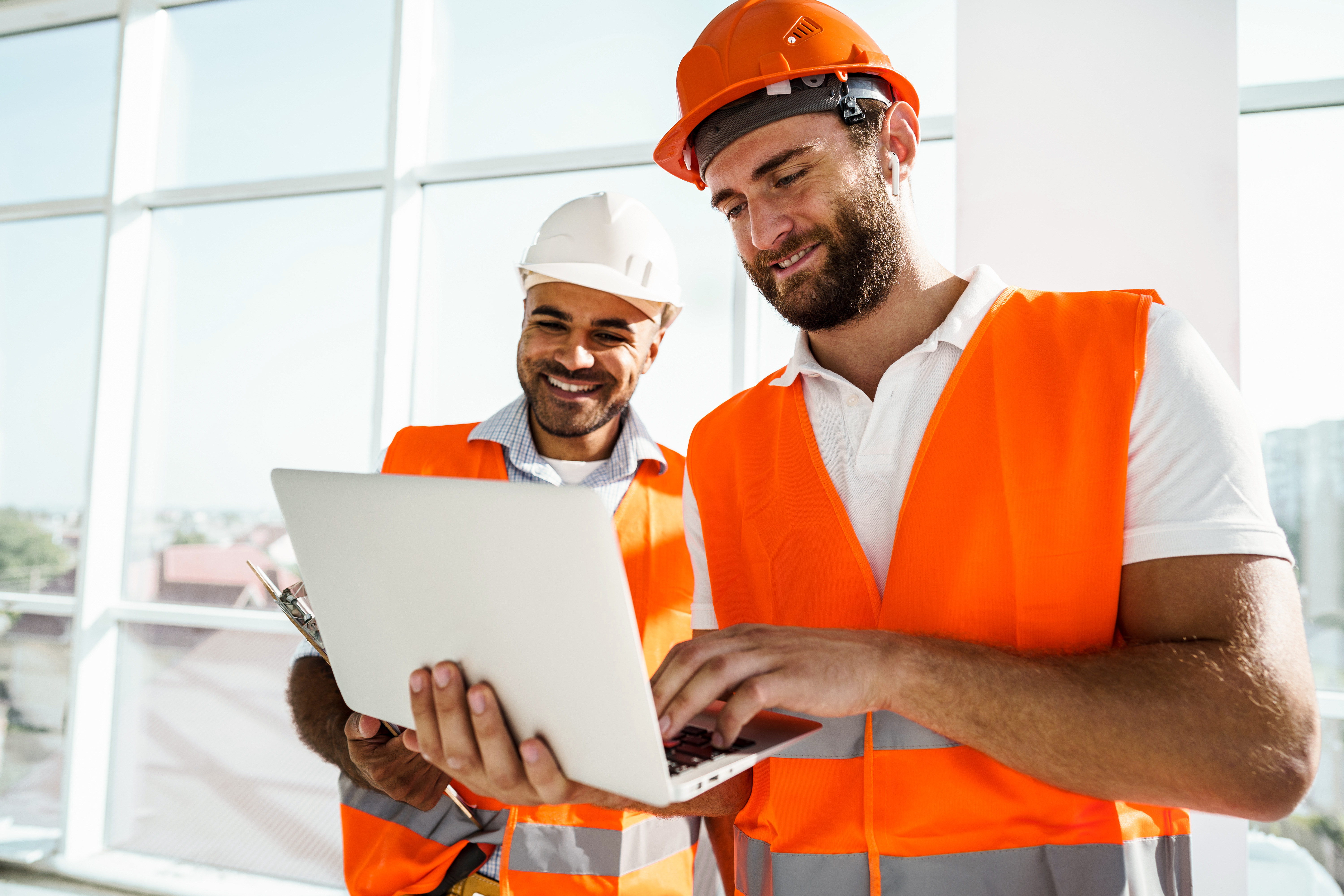 Two construction workers in safety vests and hard hats collaborate on a laptop