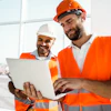 Two construction workers in safety vests and hard hats collaborate on a laptop