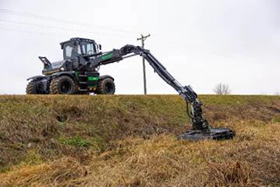 Green and black forestry harvester machine extending long mechanical arm across grassy field