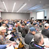 Crowded conference room with attendees seated, facing a presentation screen