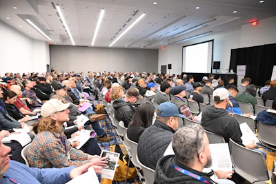 Crowded conference room with attendees seated, facing a presentation screen