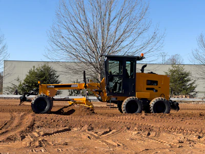 Yellow motor grader leveling dirt at construction site with bare trees and building