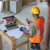 Construction worker in safety vest and hard hat reviewing plans on laptop and mobile