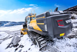 Yellow Volvo excavator on rocky snow-covered terrain with mountain backdrop