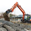 Orange excavator moving gravel at urban construction site with buildings in background