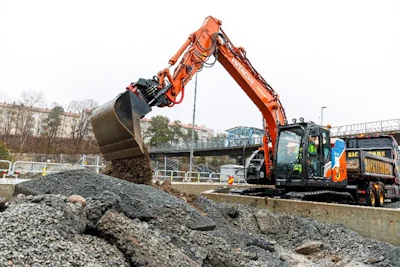 Orange excavator moving gravel at urban construction site with buildings in background