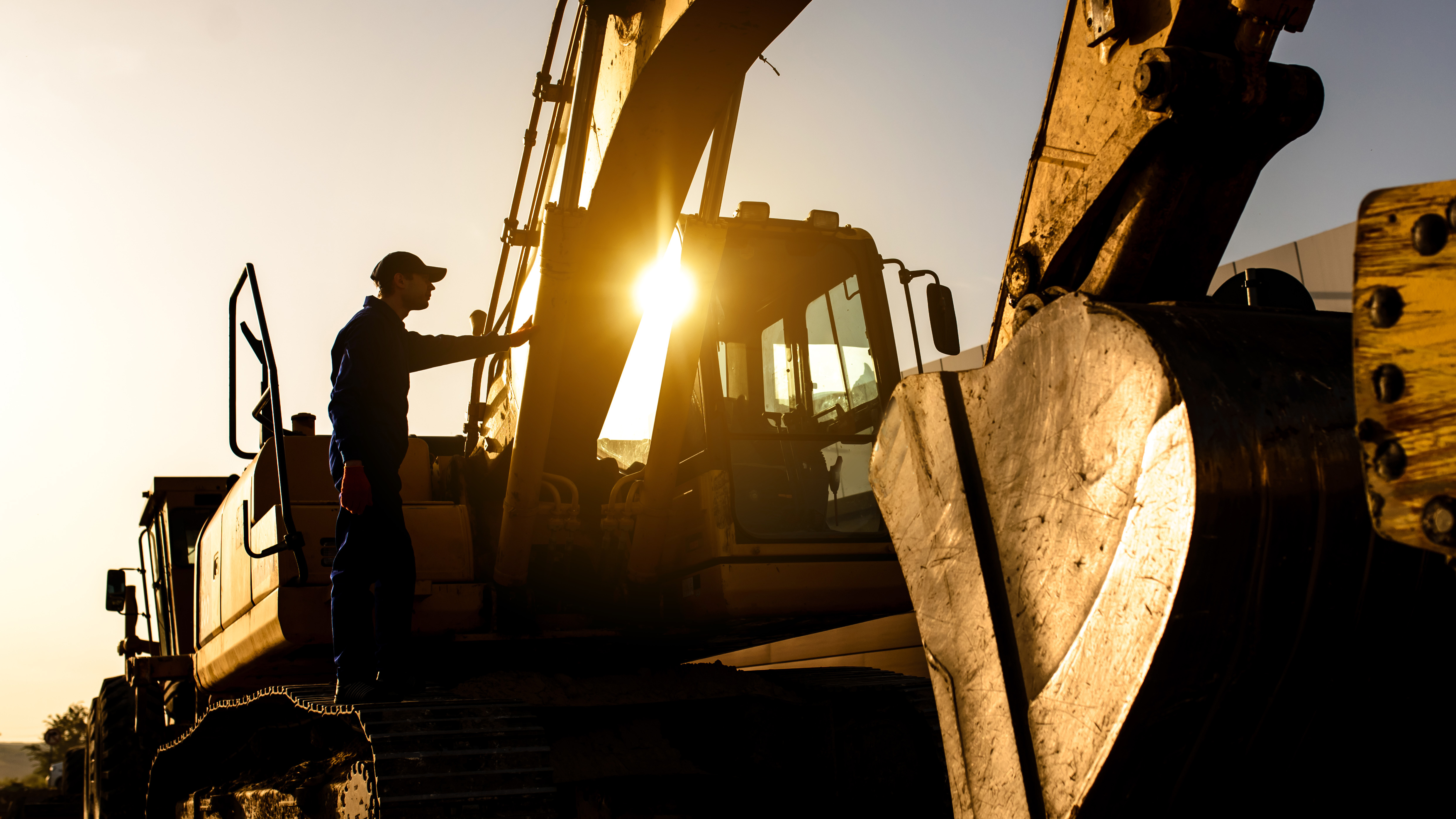 Construction worker climbing excavator at sunrise with golden backlighting