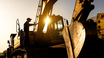 Construction worker climbing excavator at sunrise with golden backlighting