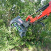 Red and gray mechanical excavator attachment clearing vegetation among green trees